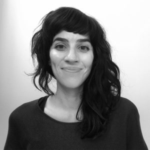 Black-and-white headshot of a person smiling gently at the camera against a plain light background. They have shoulder-length dark wavy hair with short bangs, dark eyebrows, and are wearing a dark knit sweater. The portrait is cropped from the chest up and has soft, even lighting.