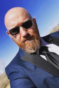 A person with a bald head and a full reddish-gray beard wears silver-framed sunglasses, a blue suit jacket with black lapels, a white dress shirt, and a black tie. The photo is taken at a slight angle outdoors against a bright blue sky, with a blurred natural landscape in the background.