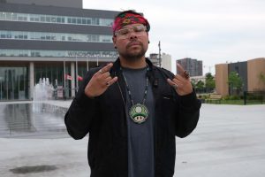 A person stands in an outdoor plaza in front of Windsor City Hall, facing the camera with both hands raised in a celebratory gesture. They are wearing a black jacket over a gray shirt, a red floral headband, clear-framed glasses, several rings, and a beaded medallion necklace featuring a green mushroom design. Fountains and Canadian flags are visible in the background beneath an overcast sky.