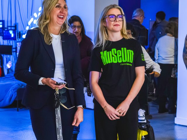 A student employee (right) wearing an MSU Museum black t-shirt chats with a visitor (left) in a dark pantsuit. Both stand in an exhibit with partially lit blue walls, screens, and wires in the bathroom.