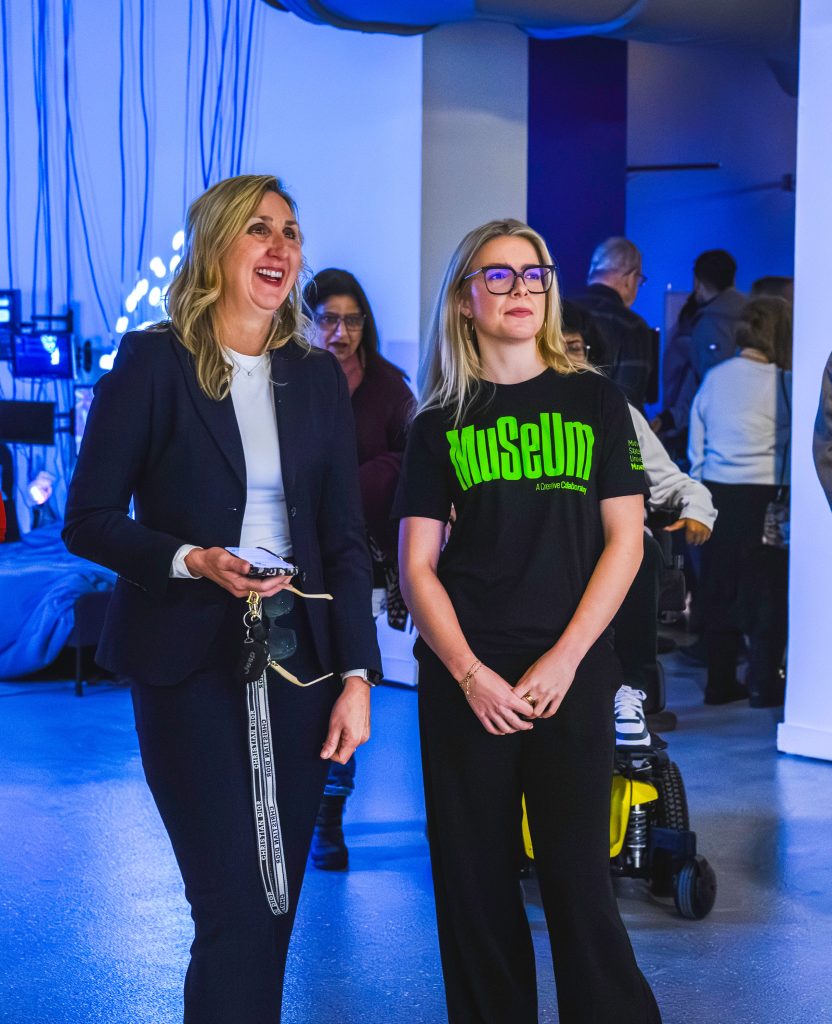 A student employee (right) wearing an MSU Museum black t-shirt chats with a visitor (left) in a dark pantsuit. Both stand in an exhibit with partially lit blue walls, screens, and wires in the bathroom.