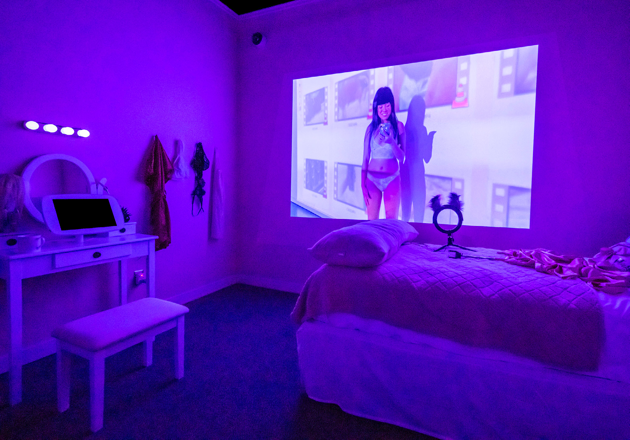 An exhibit set up like a young woman's bedroom with pink furniture, dimly lit to showcase a projection of a young woman in a sparkly bikini on the wall.
