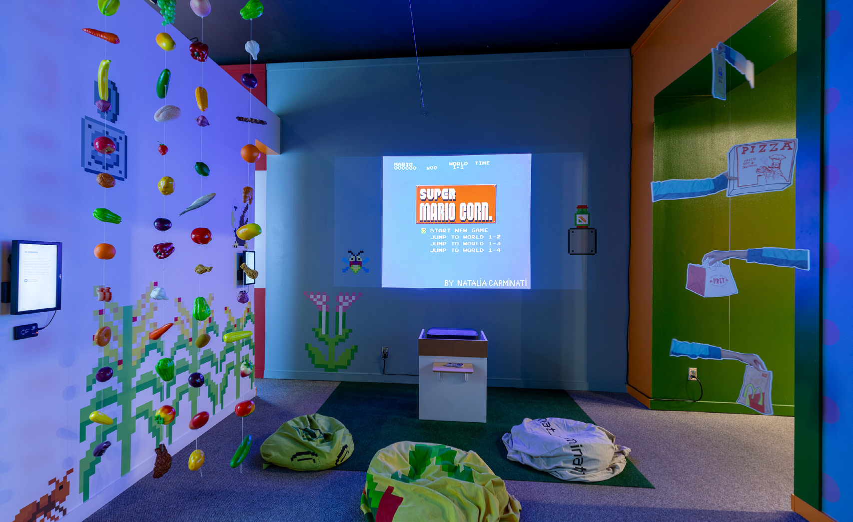 A colorful exhibit space with brightly painted walls and a projection screen displaying a game "Super Mario Corn." Bean bags sit on the floor and plastic fruit hangs on the wall to the left while paper cutouts of arms delivering food items hangs on the right.