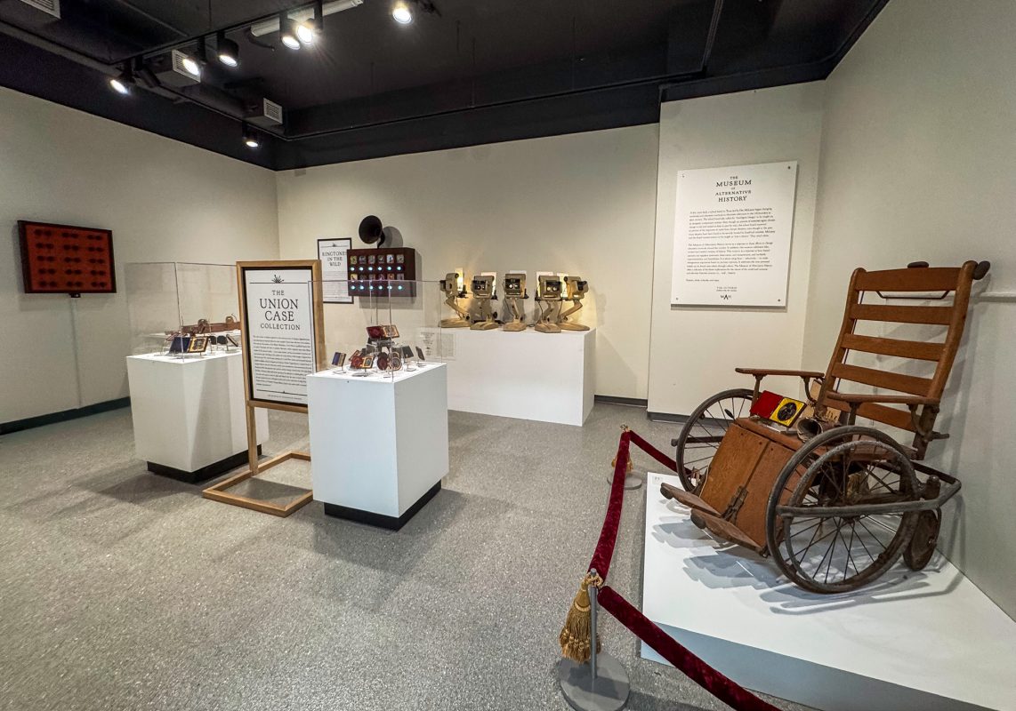 A museum gallery with white walls where artifacts sit on white pedestals including small photo portrait frames, special photo viewers, and an old wooden wheelchair.
