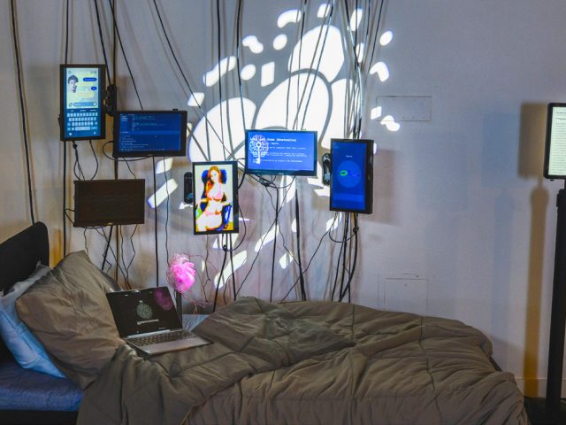 A museum exhibit featuring a bed with wrinkled grey duvet and matching pillow against a white wall covered in multiple screens and hanging wires. On the screens, various chat prompts can be seen.