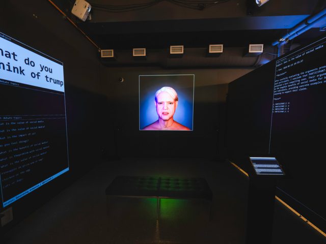 A dark museum exhibit with black walls and three projections, one per wall, with white text. Left, the projection reads "what do you think of trump," middle is a projected image of an animated older woman with short grey hair, and right is a projection of several paragraphs of white text explaining dialogue. In front of the projections sits a pedestal with an ipad and a black leather bench.