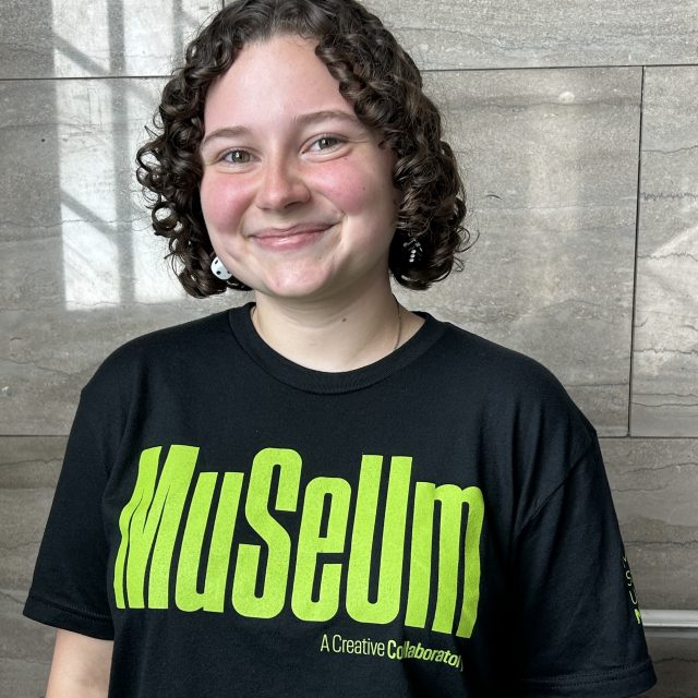 A smiling person with short, curly brown hair wears a black MSU Museum T-shirt with bright green lettering and stands in front of a gray stone wall.