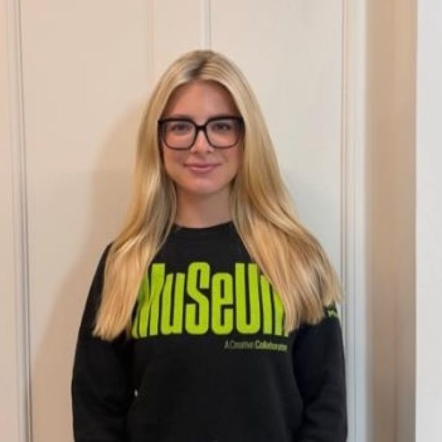A smiling person with long blonde hair and black glasses wears a black MSU Museum sweatshirt with bright green lettering and stands in front of a light-colored wall.