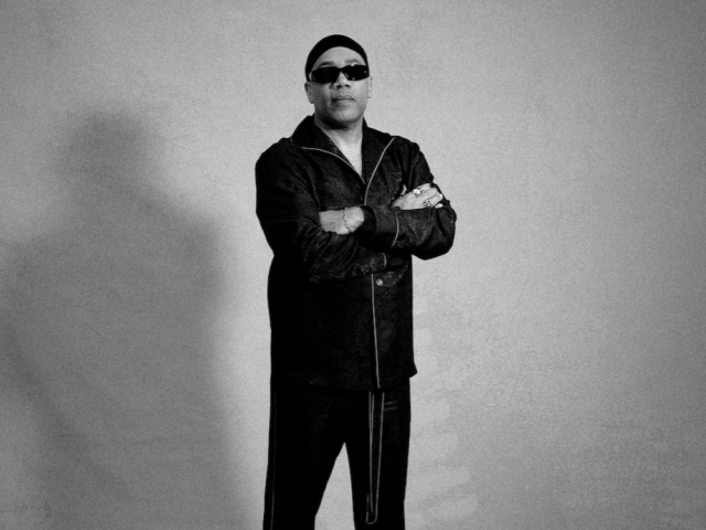 Black-and-white, full-length studio portrait of Carl Craig wearing dark sunglasses, a black jacket, black pants, and boots, standing with arms crossed against a plain gray backdrop.