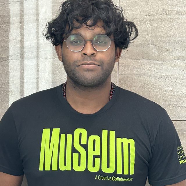 A person with curly dark hair and round glasses wears a black MSU Museum T-shirt with bright green lettering and stands in front of a gray stone wall.